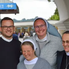 Ksiądz i trzy zakonnice uśmiechają się do zdjęcia na tle budynku z napisem "KOŚCIELE!".