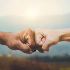 Dwie zaciśnięte pięści, jedna ciemniejsza, druga jaśniejsza, zderzają się na tle rozmytego krajobrazu. Symbol jedności i siły, jak mądrość syracha cytaty.