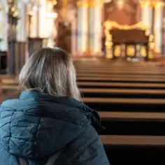 Kobieta w kościele, siedząc w ławce, zastanawia się, co to jest modlitwa. Wnętrze świątyni, pełne ozdób, sprzyja zadumie.