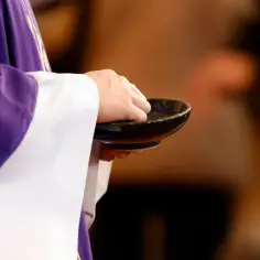 Kapłan w fioletowej dalmatyce podczas mszy świętej w środę popielcową zanurza palce w miseczce z popiołem.