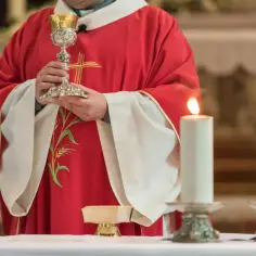 Kapłan w czerwonej szacie z haftem kłosa trzyma kielich podczas modlitwy przed przeistoczeniem. Płoną dwie świece.