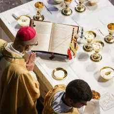 Biskup w czerwonej piusce i złotej szacie modli się nad otwartą księgą podczas drugiej modlitwy eucharystycznej. Wokół kielichy z hostiami.