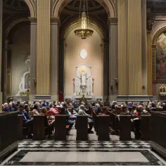 Ludzie siedzą w ławkach w kościele, przed ołtarzem z figurą Matki Boskiej.