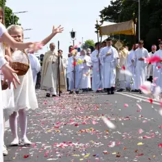 Dzieci sypią płatki kwiatów na ulicy podczas procesji Bożego Ciała. W tle widać duchownych w białych szatach niosących monstrancję.