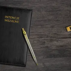 Czarny notatnik z napisem "INTENCJE MSZALNE", długopis i złoty krzyż na drewnianym tle.