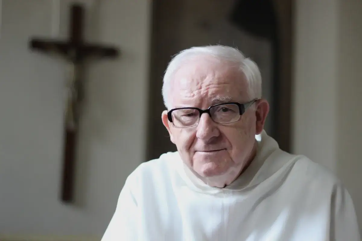 Starszy mężczyzna w okularach, w białej szacie, z krzyżem w tle. Jego spojrzenie przywodzi na myśl rozważania o dogmacie o nieomylności papieża.