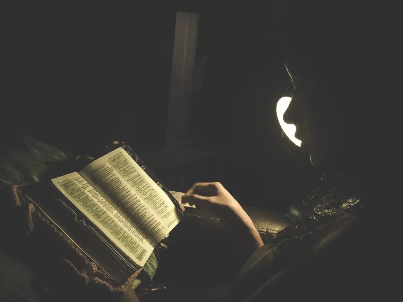 Osoba czyta Biblię przy lampce, szukając pocieszenia w trudnych chwilach, być może w obliczu oficjum za zmarłego.