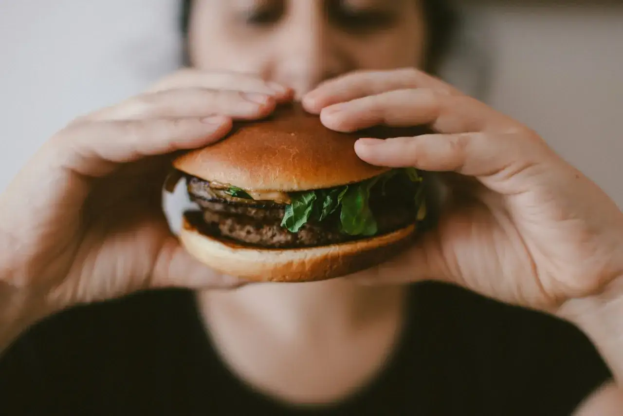 Dłonie trzymają ogromnego burgera, symbol nieumiarkowania w jedzeniu i piciu.